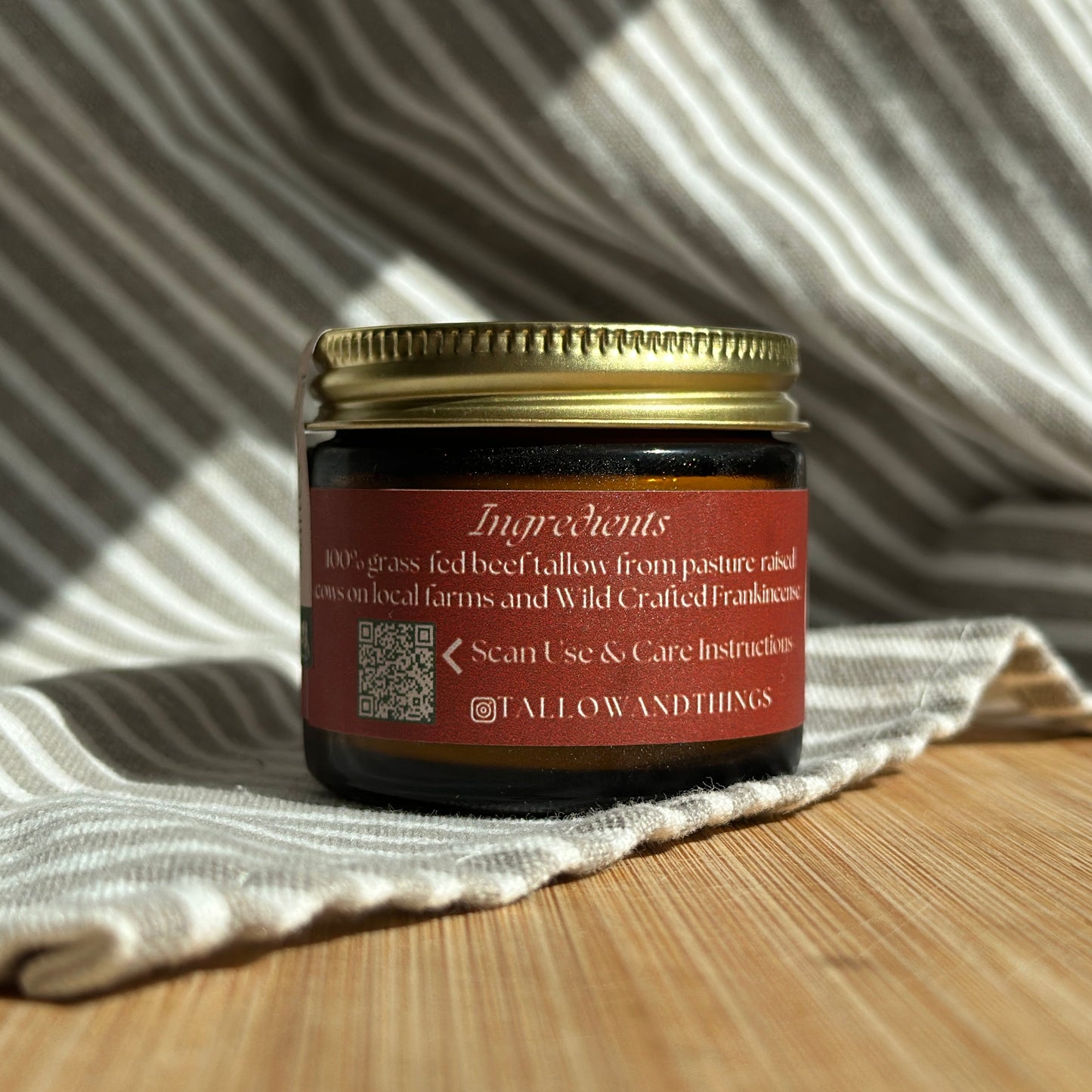 A jar of Frankincense Essential Oil Infused Skin Tallow placed on a striped cloth on a wooden surface, with a label that includes the product name and benefits.