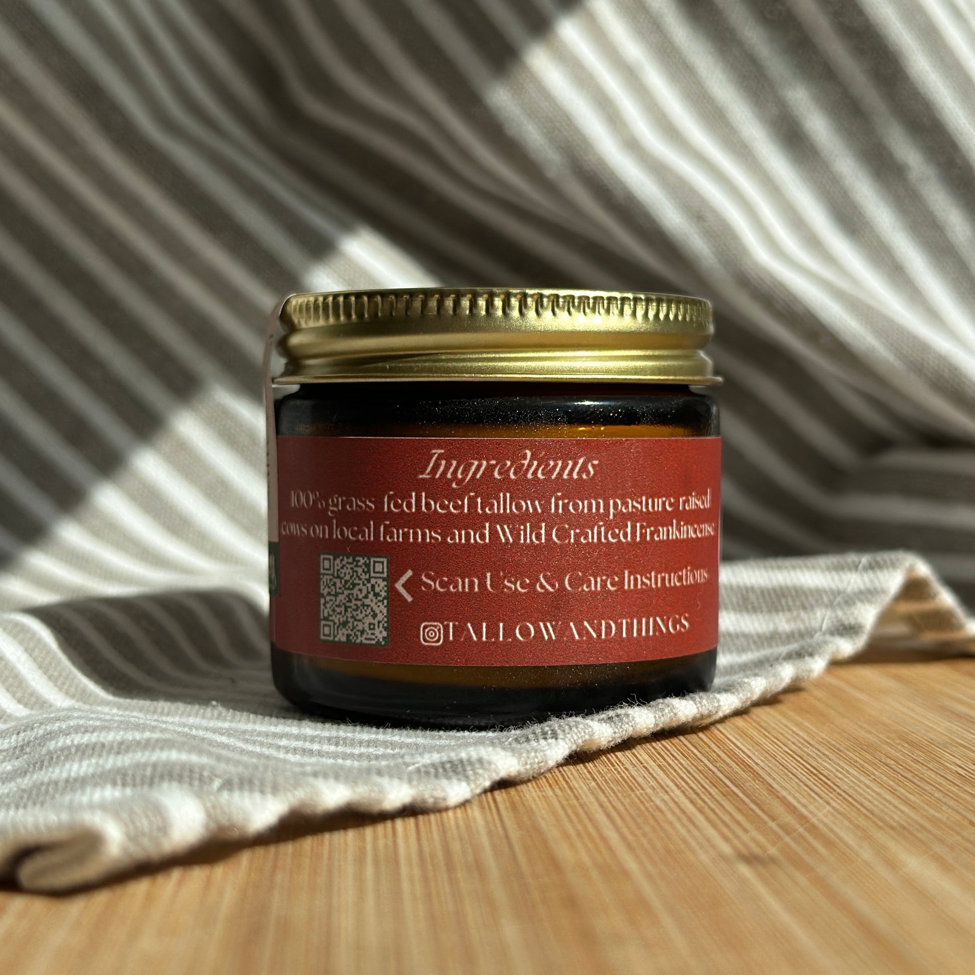 A jar of Frankincense Essential Oil Infused Skin Tallow placed on a striped cloth on a wooden surface, with a label that includes the product name and benefits.